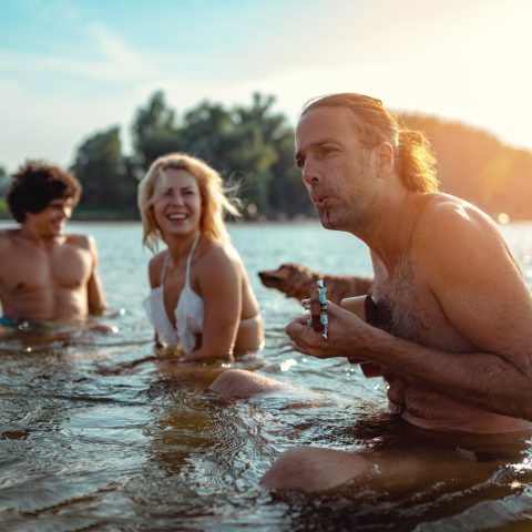 Happy young people having a great time together at the beach. They are sitting in the water and man is playing ukulele and singing. Sunset over water.